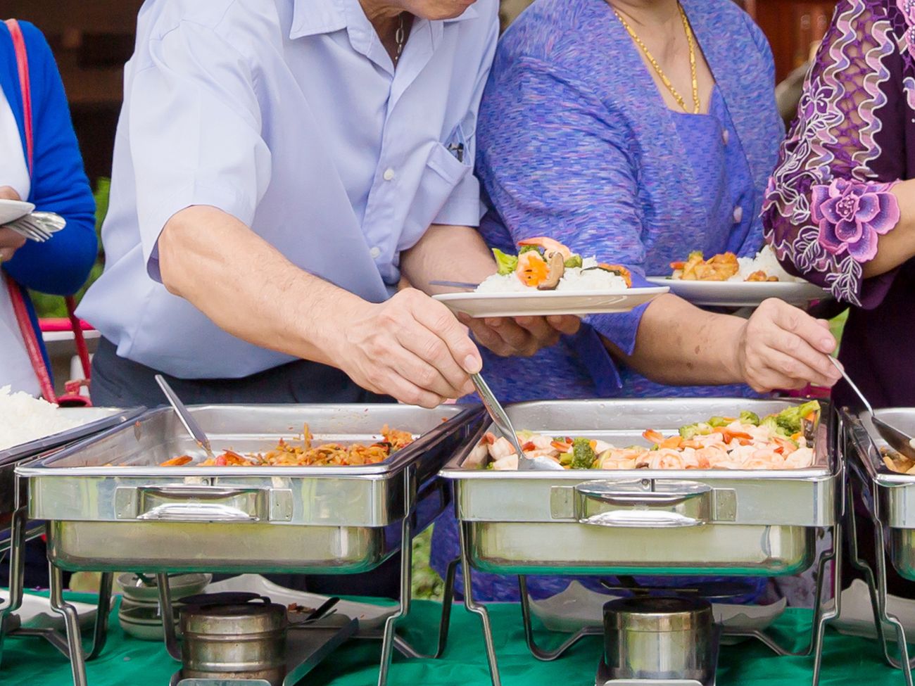 mehrere Speisen auf Warmhalteplatten, ein großer Stapel Teller und eine Warteschlage von Menschen, die sich auffüllen
