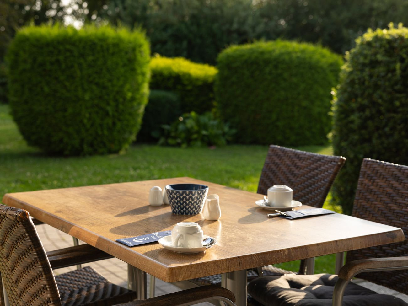 Für Frühstück gedeckter Tisch im Garten, dahinter zurechtgeschnittene Büsche