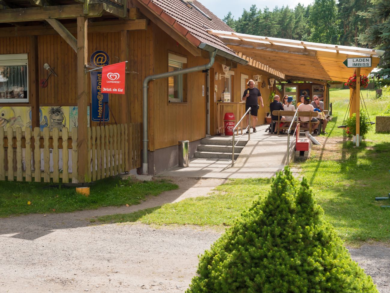 Eingangsbereich eines Kiosks, überdacht mit einem Holztisch an dem eine Gruppe von Menschen sitzt.