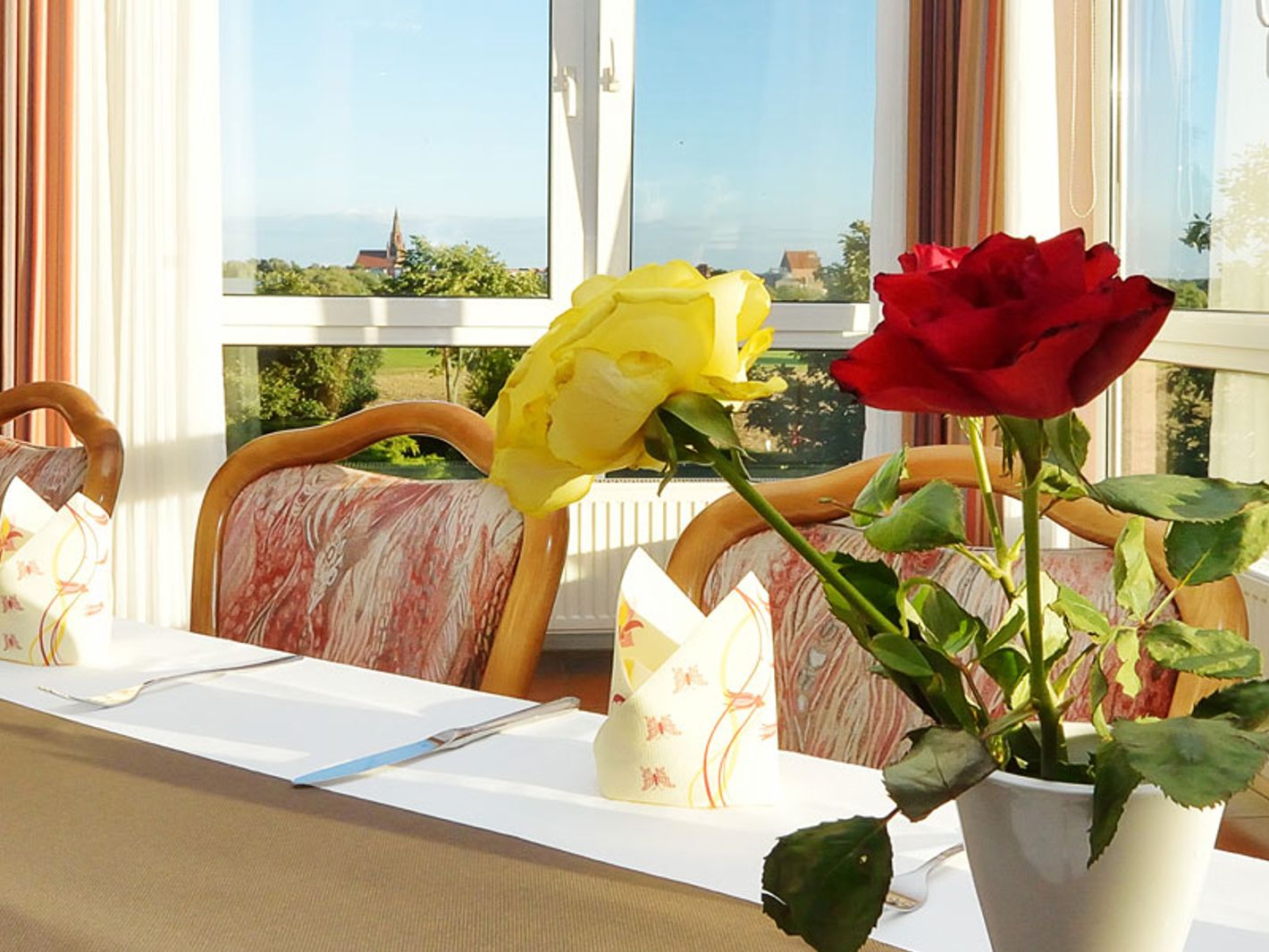 Helles, elegant eingerichtetes Esszimmer mit gedecktem Tisch. Darauf zwei Rosen – rot und gelb – in einer weißen Vase. Gemusterte Stühle, gefaltete Servietten und Pflanzen vor großen Fenstern schaffen eine warme, einladende Atmosphäre.