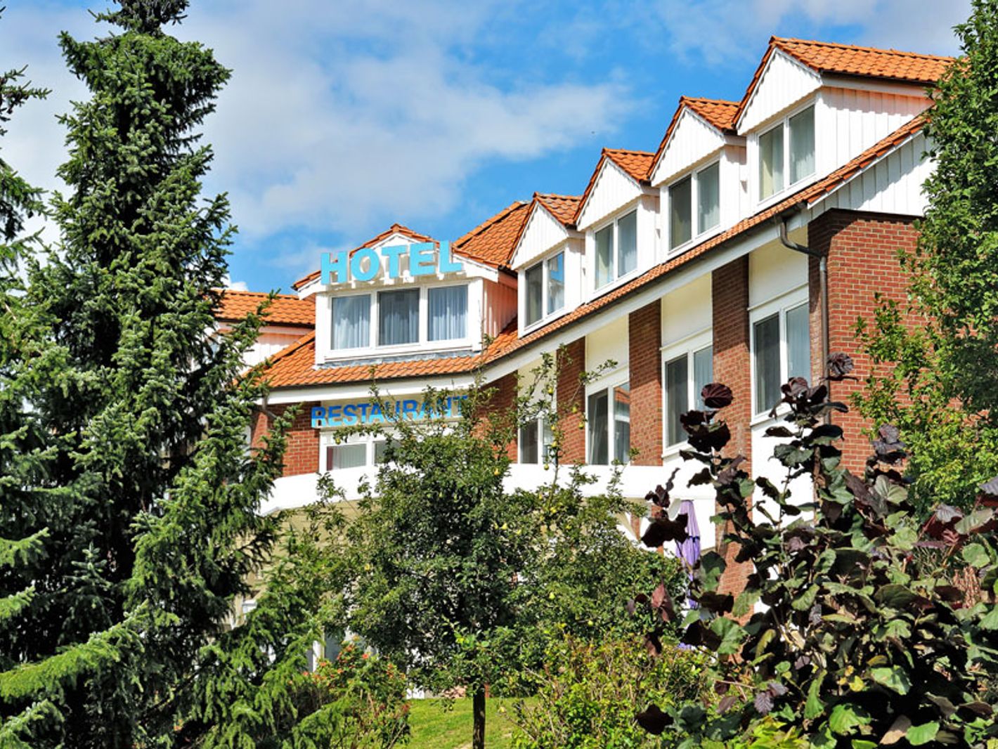 Außenansicht des Hotelgebäudes hinter Nadelbäumen unter blauem Himmel mit ein paar Quellwolken