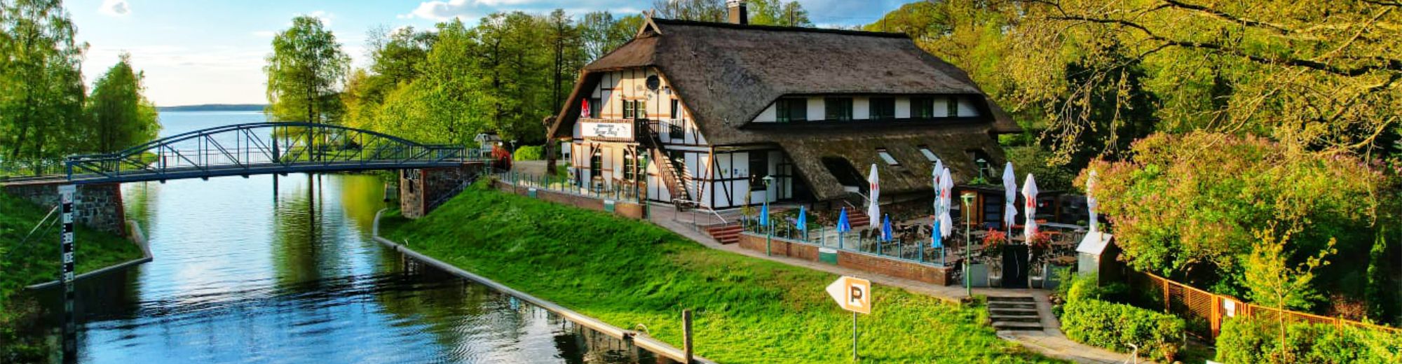 Reetgedecktes Gebäude am Wasser, direkt daneben eine Fußgängerbrücke