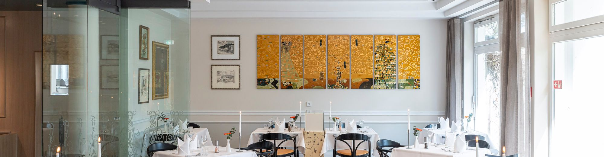 heller Gastraum des Restaurants mit bodentiefen Fenster, Luster an der Decke, Holzfussboden. Die Tische sind für ein 3-gängiges Menü eingedeckt, an den Wänden hängen Jugenstilgemälde.