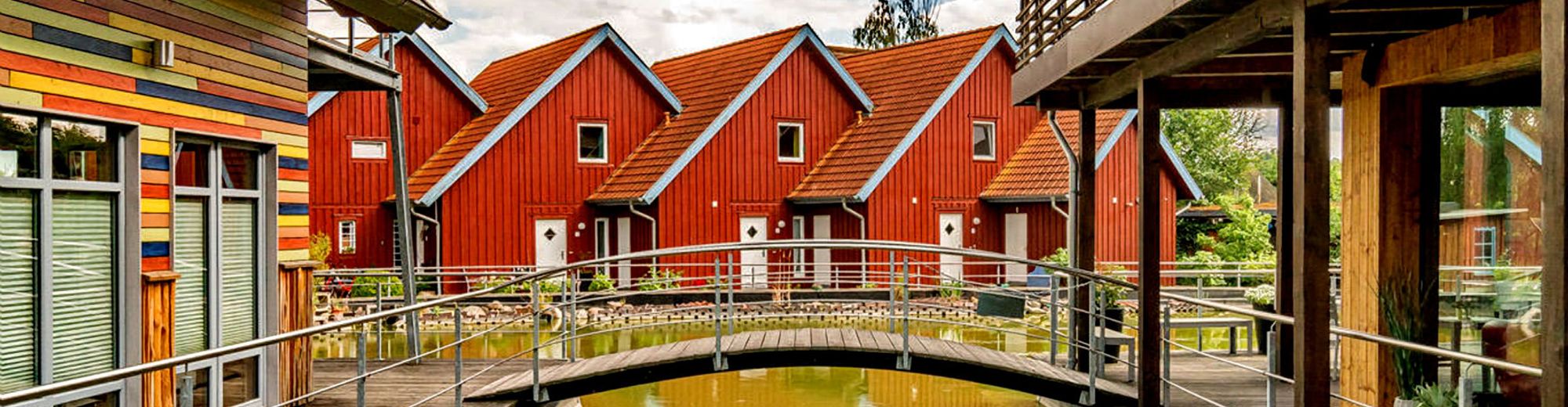 Naturteich mit Brücke, dahinter Ferienhäuser mit roter Holzfassade
