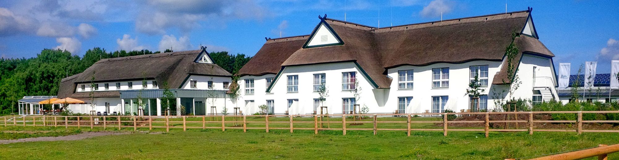 Außenansicht des Hotels mit weißer Fassade und Reetdach, davor eine mit Holz eingezäunte Pferdekoppel