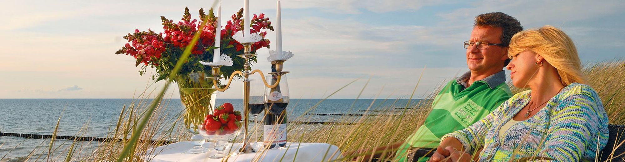 ein Paar am Strand vor einem kleinen Tischchen mit Wein und Erdbeeren und einem roten Blumenstrauß