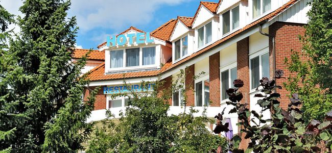 Außenansicht des Hotelgebäudes hinter Nadelbäumen unter blauem Himmel mit ein paar Quellwolken