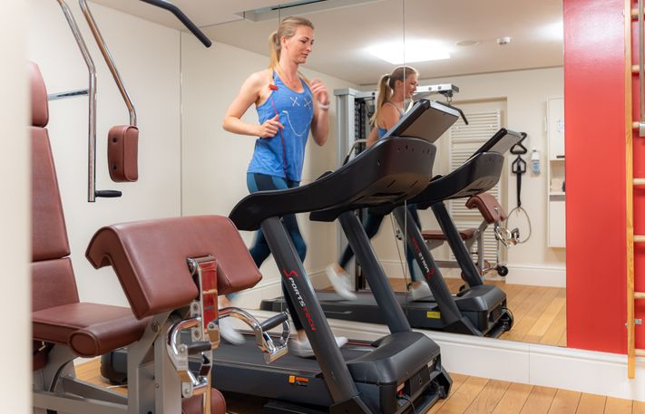 Fitnessraum mit Fitnessgeräten und Spiegelwand. Eine Frau läuft auf einem Laufband.