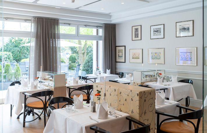 Lichtdurchfluteter Gastraum mit bodentiefen Fenstern, eingedeckte Tische mit schwarzem Mobiliar.