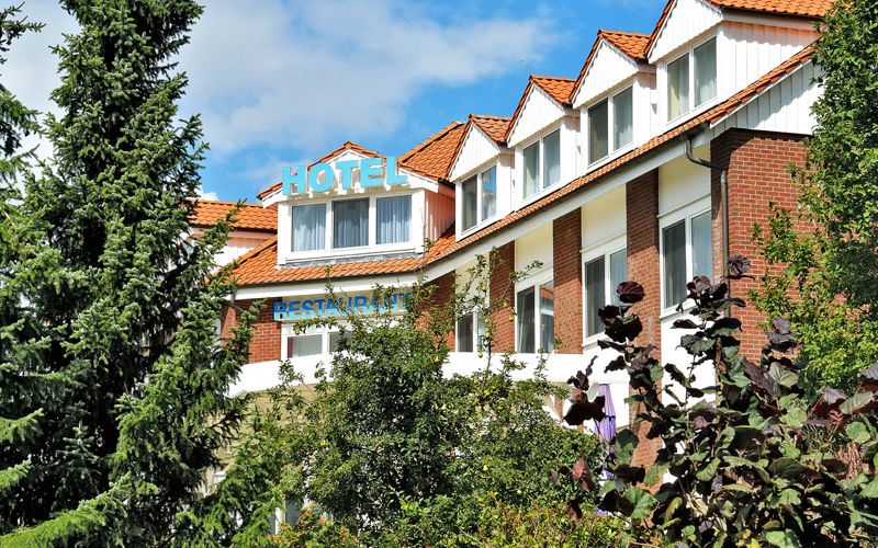 Außenansicht des Hotelgebäudes hinter Nadelbäumen unter blauem Himmel mit ein paar Quellwolken