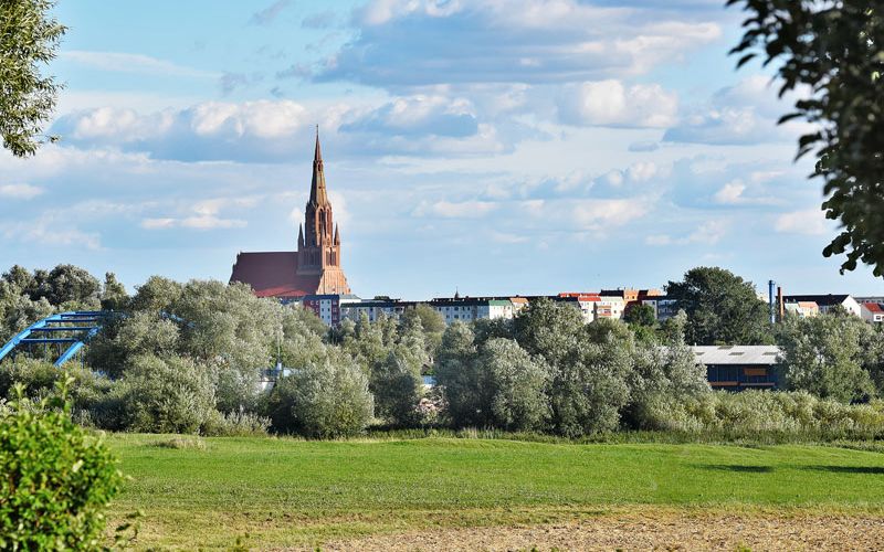 grüne Wiese mit kurzem Gras, im Hintergrund begrünte Bäume, eine Brücke mit blauem Metallgeländer und die Kirche von Demmin
